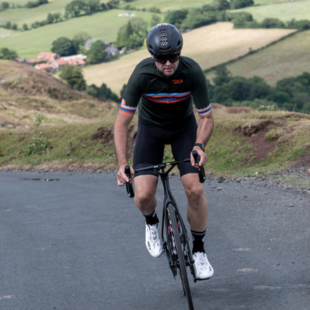 Male rider cycling up a hill out of the saddle riding towards the camera wearing men's Groove jersey