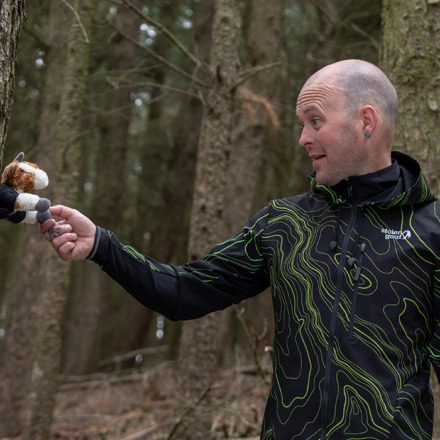 Man wearing MTB Enduro jacket in woods shaking hands with Baby Scruff small plush goat toy