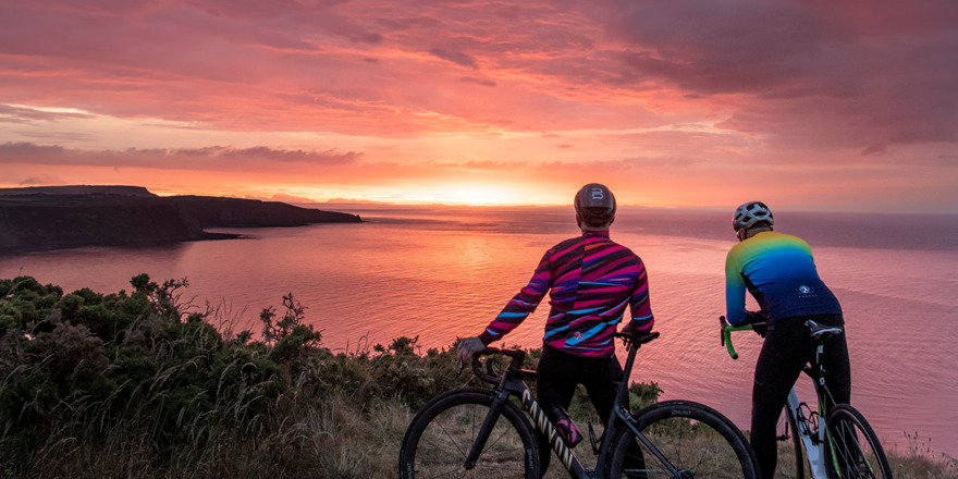 Two cyclists wearing men's AW22 kit standing on the edge of a cliff looking out at a sunset over the sea