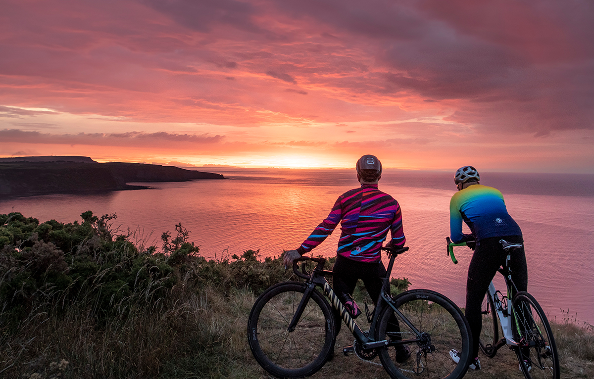 Two cyclists wearing men's AW22 kit standing on the edge of a cliff looking out at a sunset over the sea