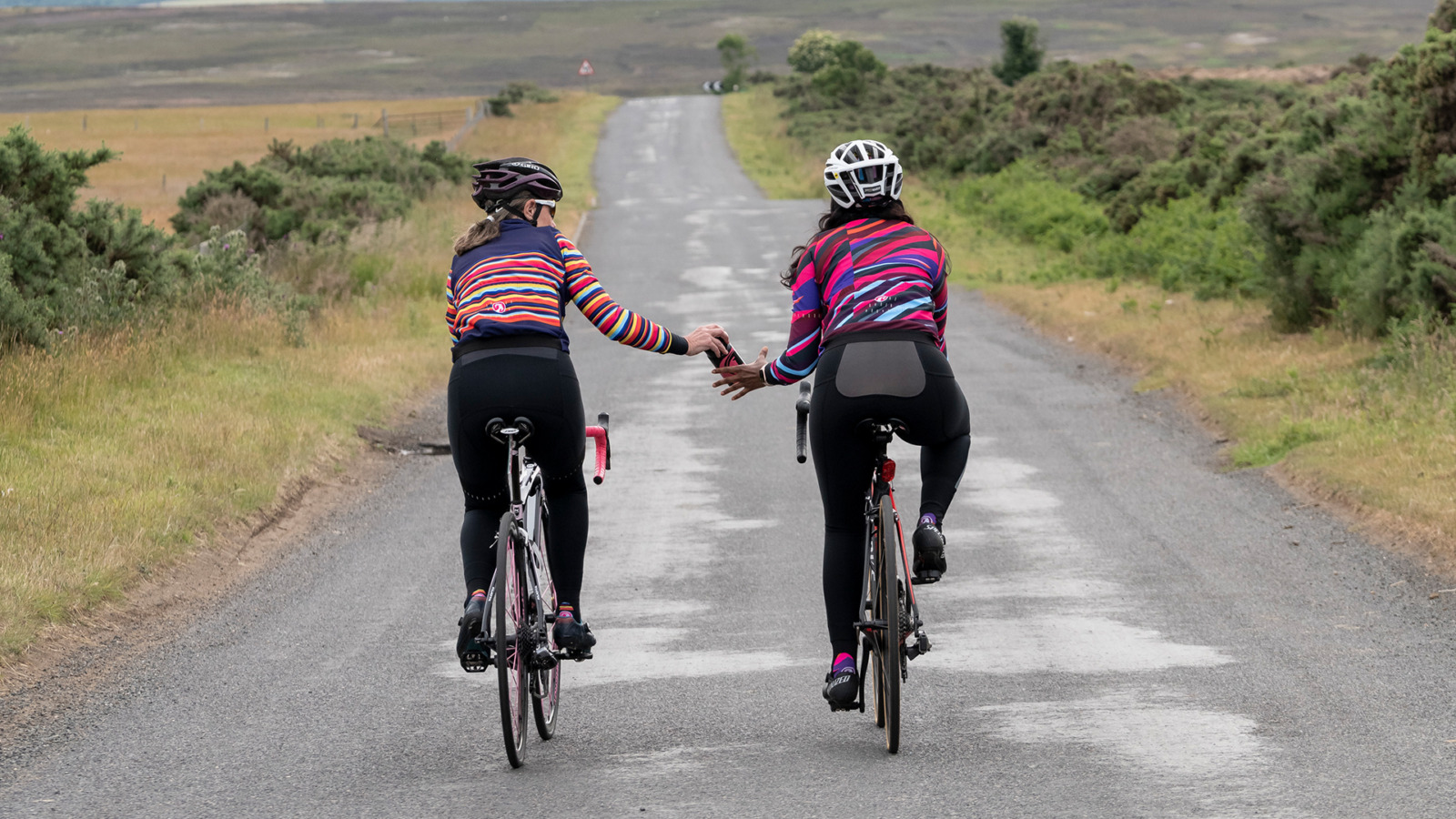 Two cyclists in women's Alpine jackets riding down a road passing a water bottle