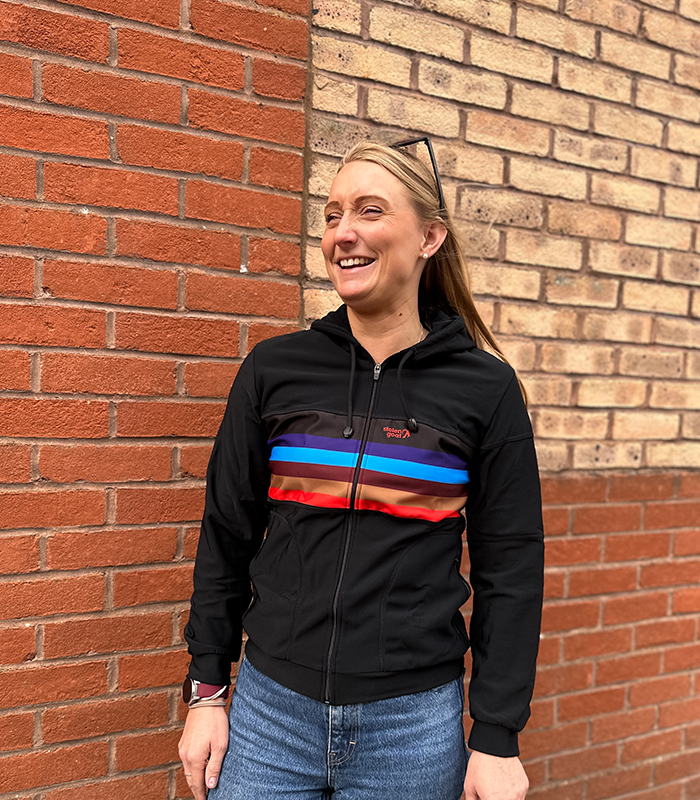 Woman wearing Manta Kiko hoodie, black hoodie with red, gold, burgundy and blue block stripe across the centre of the chest.