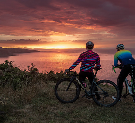 two men wearing sliver and fluid alpine jackets at the top of a cliff watching the sunset over the sea