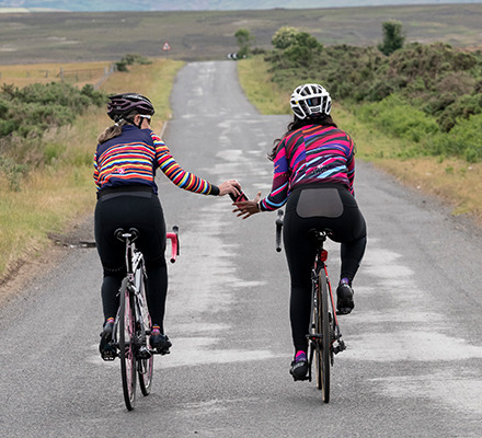 two women riding next to each other passing a water bottle wearing doofs and sliver alpine bodyline jackets