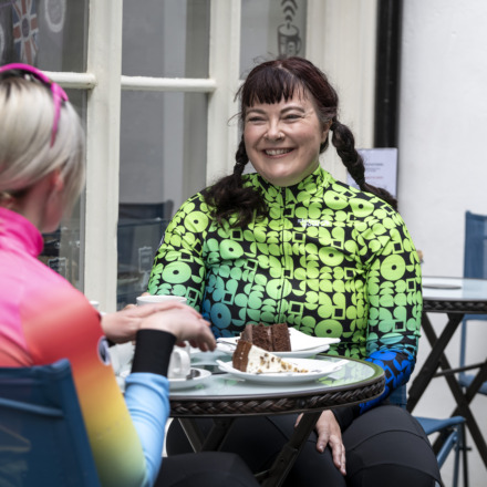 Two women cyclists talking at a cafe stop wearing Stolen Goat Kiko jerseys
