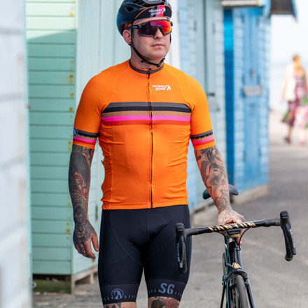 Man standing in front of beach huts pushing his bike while wearing orange quadrant cycling jersey