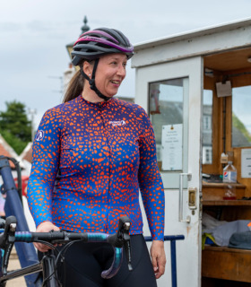 Woman wearing blue and orange print Strutter jersey standing next to her bike smiling