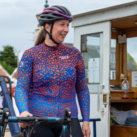 Woman wearing blue and orange print Strutter jersey standing next to her bike smiling